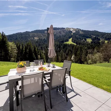 Ein schöner Außenbereich mit einem Tisch und Stühlen auf einer Wiese. Im Hintergrund sind bewaldete Hügel und ein blauer Himmel zu sehen.