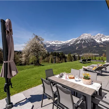 Ein schöner Außenbereich mit einem Tisch und Stühlen, umgeben von grüner Landschaft und Bergen im Hintergrund. Die Sonne scheint auf die Terrasse, die mit einem Sonnenschirm ausgestattet ist.