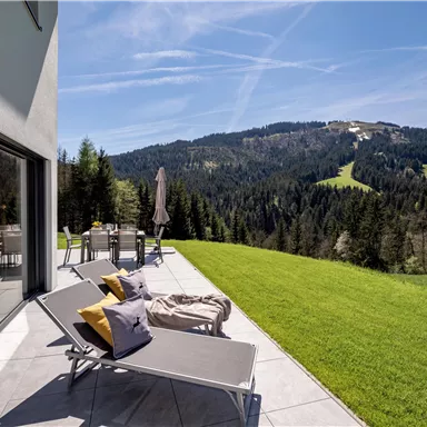 Eine moderne Terrasse mit Liegen und bunten Kissen bietet einen Blick auf grüne Hügel und Wälder. Der Himmel ist klar und blau, ideal für entspannte Tage in der Natur.