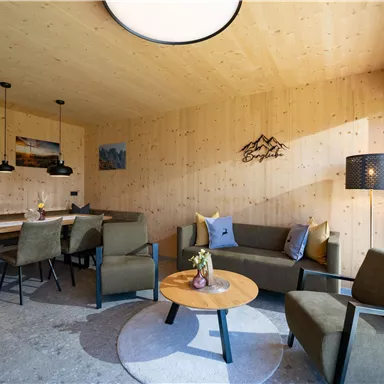 Ein modernes Wohnzimmer mit Holzvertäfelung und einem runden Tisch. Gemütliche Sitzmöbel und große Fenster sorgen für eine helle Atmosphäre.