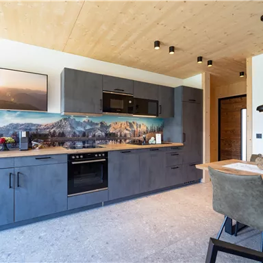 Eine moderne Küche mit grauen Schränken und Holzdecke. Ein Esstisch mit Stühlen steht neben der Küchenzeile.