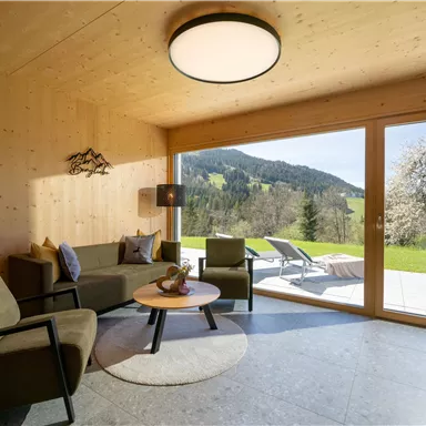Ein modernes Wohnzimmer mit Holzwänden und großen Fenstern. Der Blick geht auf eine grüne Landschaft mit Bergen im Hintergrund.
