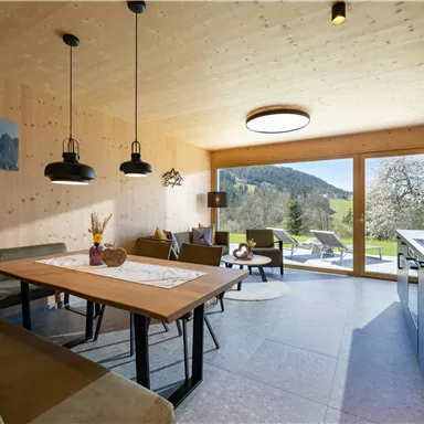 Eine moderne Wohnküche mit Holzwänden und großen Fenstern. Der Raum bietet einen Blick ins Grüne und ist gemütlich eingerichtet.