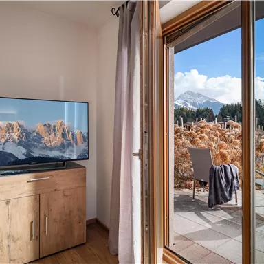 A cozy room with a television and a view of the mountains. Outside is a terrace with chairs and a beautiful landscape.
