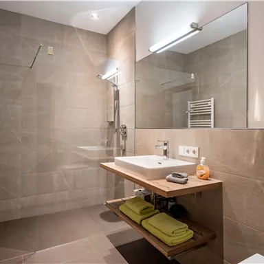 A modern bathroom with a large shower and a bright sink. The walls are painted in neutral tones and there are towels on a wooden shelf.