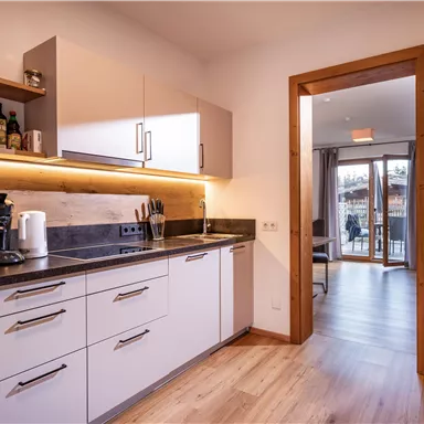 A modern kitchen with a wooden floor and bright lighting. Through the open door, a living area with access to the balcony is visible.