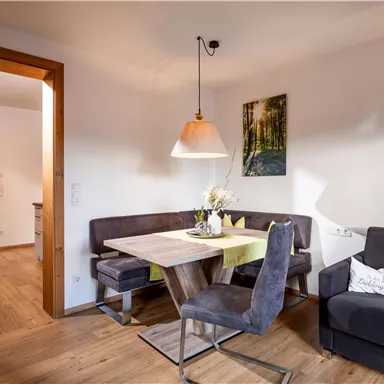 A cozy living area with a dining table, chairs, and a sofa corner. A picture hangs on the wall, and the light is pleasant and warm.