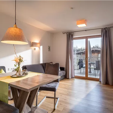 A modern living room with a dining table and a cozy couch. Large windows provide a view of the terrace outside.