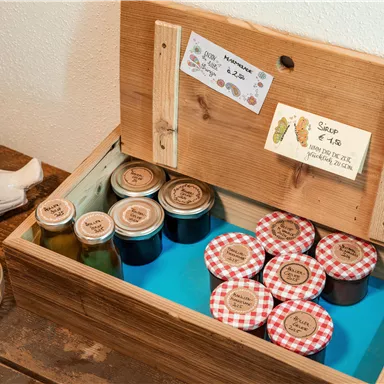 A wooden box with various jars and jam with checkered lids. It also contains small signs with prices.