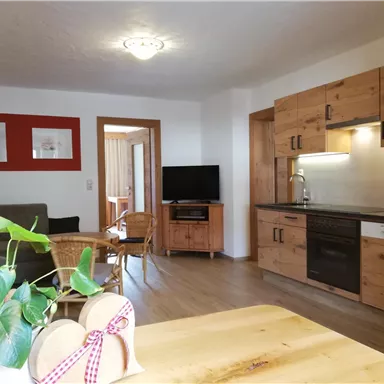 A modern kitchen with wooden furniture and a dining table. In the background, there is a cozy living area and a television.