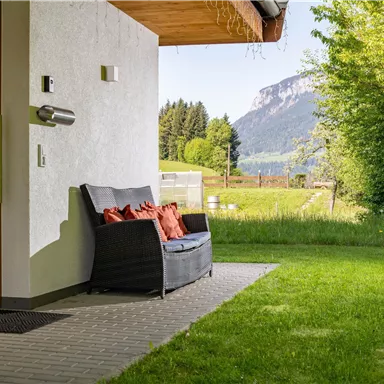 A cozy seating area with cushions on a terrace. In the background, green meadows and hilly mountains can be seen.