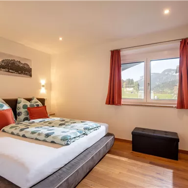 A cozy bedroom with a double bed and colorful pillows. Large windows offer a view of the surrounding landscape.