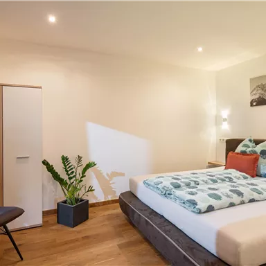 A modern bedroom with a large bed, colorful pillows, and a floor lamp. In the room, there is a wardrobe and a plant.