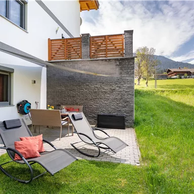 A relaxed terrace with loungers and a table in a green environment. In the background, meadows and mountains can be seen.