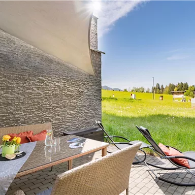An inviting terrace with a table and chairs, surrounded by a green meadow and mountains in the background. The sky is clear and sunny, perfect for relaxing outdoors.