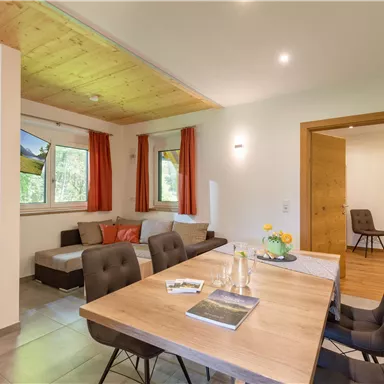 A modern kitchen living area with a wooden table and comfortable chairs. In the background, there are large windows with a view and cozy seating.