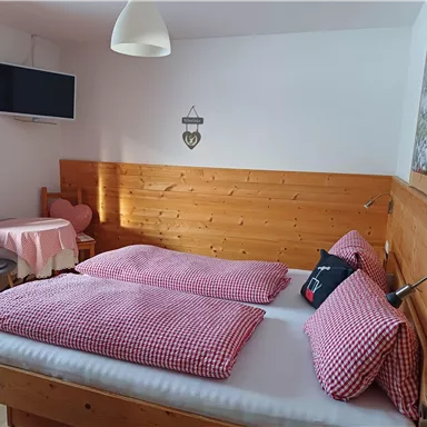 Ein gemütliches Schlafzimmer mit zwei Betten und roten Kissen. Es gibt einen kleinen Tisch, einen Stuhl und einen Fernseher an der Wand.