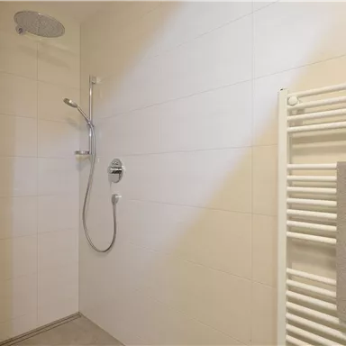 Eine moderne Dusche mit weißen Fliesen und einem Handtuchhalter. Der Duschkopf hängt an der Wand und sorgt für eine einfache Anwendung.
