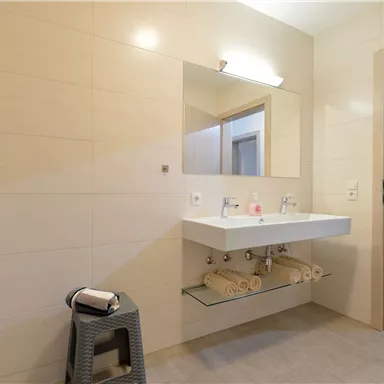 Ein modernes Badezimmer mit einem großen Spiegel und einem hellen Waschtisch. Handtücher und Rollen auf einem Regal vervollständigen die entspannende Atmosphäre.