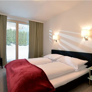 Ein modernes Schlafzimmer mit einem Doppelbett und weißen Bettdecken. Der Raum hat große Fenster mit grünen Vorhängen und einen Fernseher an der Wand.