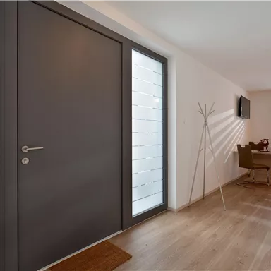 Ein moderner Eingangsbereich mit einer großen, grauen Tür und viel natürlichem Licht. Rechts befindet sich ein Essbereich mit einem Tisch und Stühlen.