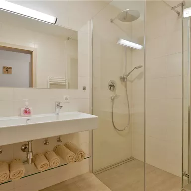 Ein modernes Badezimmer mit einer begehbaren Dusche und einem schlichten Waschbecken. Auf dem Regal liegen mehrere frisch gefaltete Handtücher.