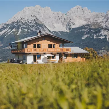 A beautiful house in the mountains surrounded by green grass. In the background, impressive mountain peaks can be seen.