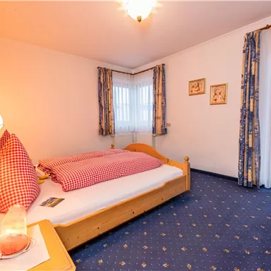 Ein gemütliches Schlafzimmer mit einem einzelnen Bett und rot-weißem Bettzeug. Die Wände sind hell, und es gibt Vorhänge mit Blumenmustern sowie eine warme Beleuchtung.