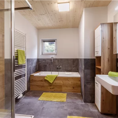 A modern bathroom with wood and stone accents. There is a shower, a bathtub, and fresh green towels.