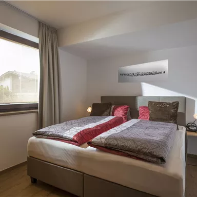 Ein modernes Schlafzimmer mit zwei Betten, dekoriert in neutralen Tönen und roten Akzenten. Ein großes Fenster sorgt für viel Tageslicht.