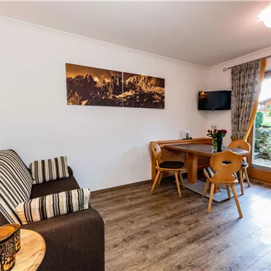 Ein gemütlicher Wohnraum mit einem Sofa, einem Essbereich und einem Fernseher. Große Fenster bieten einen Blick ins Freie und sorgen für viel Licht.