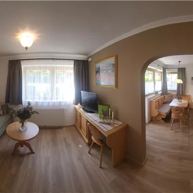 Ein gemütliches Wohnzimmer mit einem grünen Sofa, einem Couchtisch und Fenster mit hellem Licht. Im Hintergrund ist ein Essbereich mit einem Holzstuhl und Tisch sichtbar.