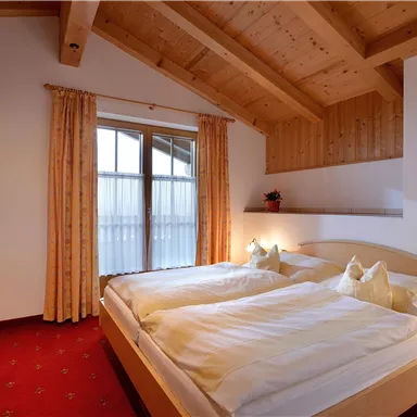 Ein gemütliches Schlafzimmer mit zwei Betten und Holzdecke. Große Fenster lassen viel Licht herein.