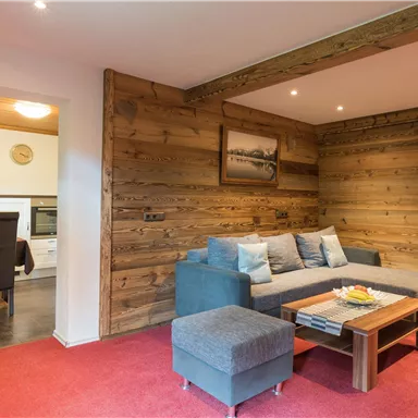 Ein gemütliches Wohnzimmer mit einer Holzverkleidung und einem roten Teppich. Im Hintergrund ist ein Essbereich mit einem Tisch und Stühlen sichtbar.