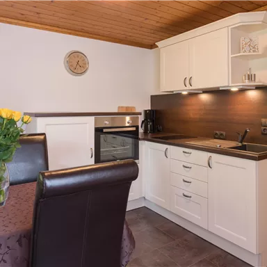 Eine moderne Küche mit weißen Schränken und einer dunklen Arbeitsplatte. Am Tisch steht eine Vase mit frischen Blumen und eine Uhr an der Wand.