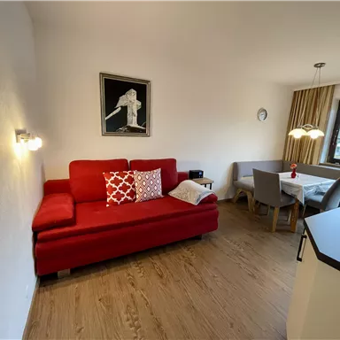 Ein gemütliches Wohnzimmer mit einem roten Sofa und einem Essbereich. Die Wände sind hell und das Licht ist warm, was eine einladende Atmosphäre schafft.