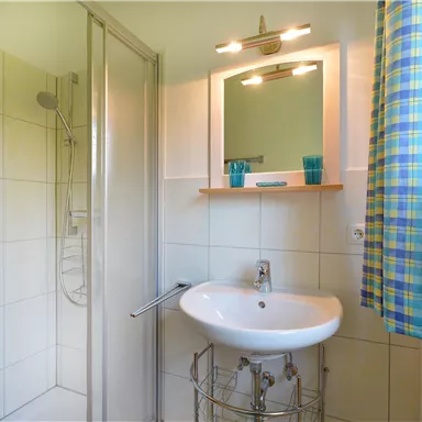 Ein modernes Badezimmer mit einer Dusche und einem Waschbecken. Es gibt ein Fenster mit einem karierten Vorhang und eine kleine Pflanze.