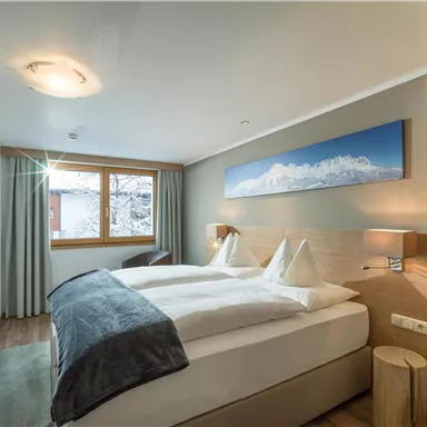 Ein modernes Schlafzimmer mit einem großen Bett und zwei Kissen. Große Fenster lassen viel Tageslicht herein und zeigen eine schöne Aussicht.