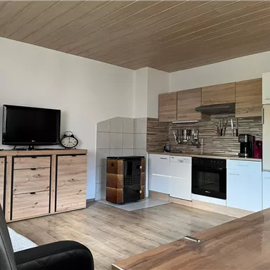 A modern kitchen with wooden cabinets and a stove. The space is bright and friendly, with a television and a dining area.