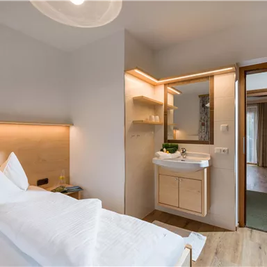 A modern bedroom with a double bed and a small wash area. There is warm light and access to an adjoining room.