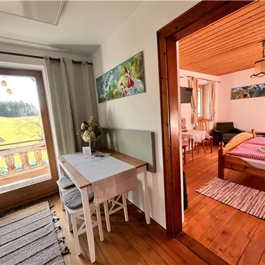 Ein gemütliches Zimmer mit Holzfußboden und einem Tisch. Durch das Fenster blickt man auf die grüne Landschaft.