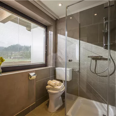 Ein modernes Badezimmer mit einer Glasdusche und einem eleganten Waschbecken. Durch das Fenster sichtbar ist eine grüne Landschaft im Hintergrund.