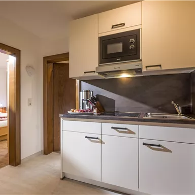 Eine moderne Küche mit weißen Schränken und eingebautem Ofen. Im Hintergrund ist ein Zugang zu einem Schlafzimmer zu sehen.