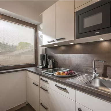 Eine moderne Küche mit hellen Schränken und einem Fenster. Auf der Arbeitsplatte liegen Obst und eine Kaffeemaschine ist sichtbar.