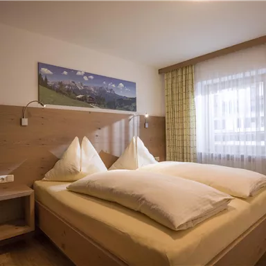 Ein gemütliches Schlafzimmer mit einem Doppelbett und zwei großen Kissen. An der Wand hängt ein Bild der Berge, während das Fenster viel Tageslicht hereinlässt.