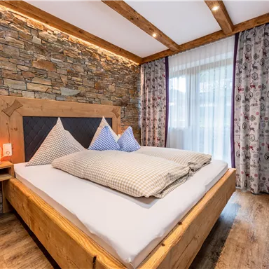 Ein modernes Schlafzimmer mit einem großen Holzbett und gemütlichen Kissen. Die Wände sind mit Stein und einem elegantem Vorhang dekoriert, während ein Fernseher an der Wand montiert ist.
