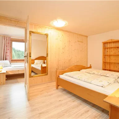 A simple bedroom with wooden paneling and a large window. There are two beds, a bedside table, and a wardrobe.