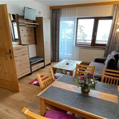 A cozy living space with wooden floors and modern furniture. There is a dining table with flowers, a television, and large windows that let in plenty of light.