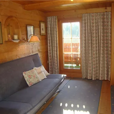 Ein gemütliches Wohnzimmer mit Holzverkleidung und einem grauen Sofa. Große Fenster mit Vorhängen lassen viel Licht herein.