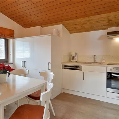 A modern kitchen with a dining table and white furniture. The floor is bright, and the wooden ceiling creates a warm atmosphere.